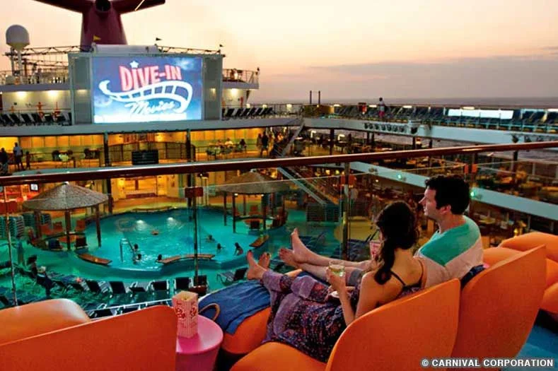 A couple sits on an upper deck of a carnival cruise ship, watching a movie on a giant video screen