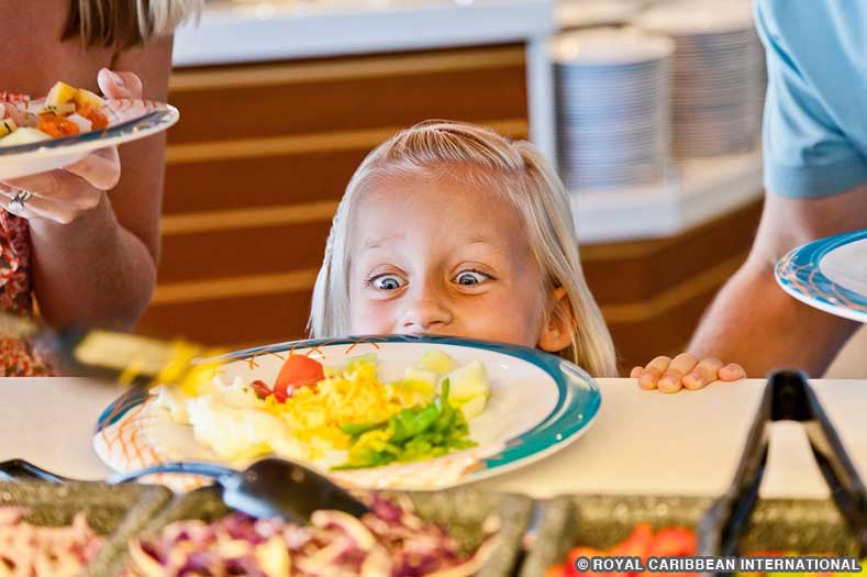A young child excitedly peeks over the counter of a buffet to view a plate of food