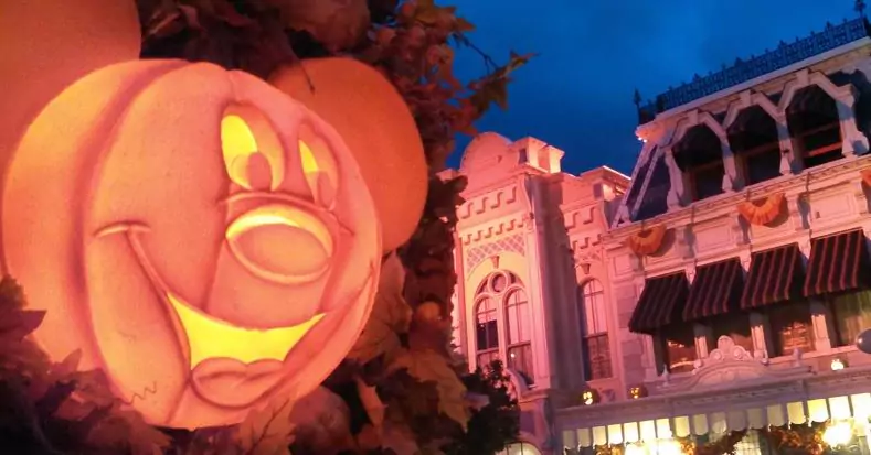 An orange Mickey Mouse jack-o-lantern glows in front of lit buildings on Main Street USA
