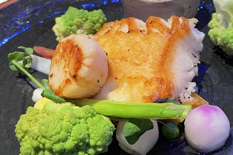 Disney Cruise Food – Chilean Sea Bass Disney Cruise Food - Chilean Sea Bass served with a seared scallop and vibrantly-colored vegetables on a dark plate.