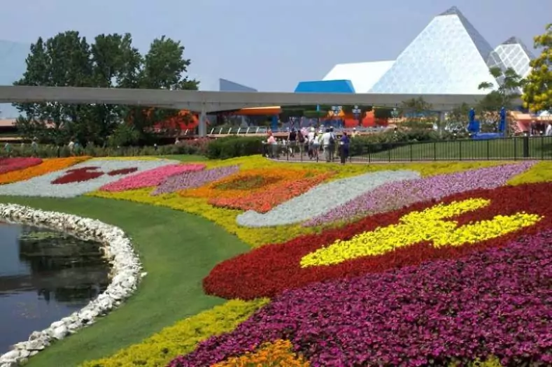 Colorful flower patches line a grassy hillside leading to a waterway at EPCOT. Some of the flowers are arranged into patterns such as a Mickey Mouse silhouette.