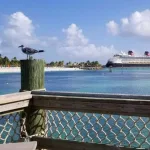 Castaway Cay - Disney's Private Island -- A Disney cruise ship is seen across crystal waters from a wood dock with net railing. A bird sits on the post.