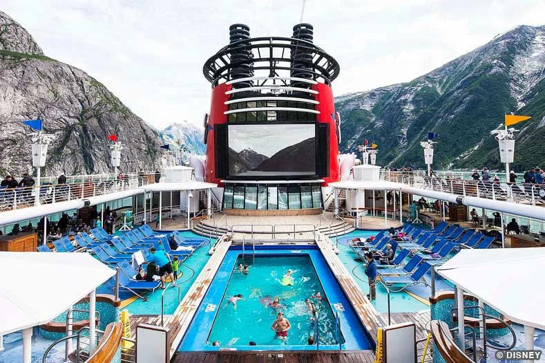 People swim in a pool on the deck of the Disney Wonder while the ship sails through a fjord