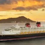 Last Minute Disney Cruise - the Disney Wonder Cruise Ship sails in front of a Hawaiian Mountain, which is silhouetted against an orange sky.