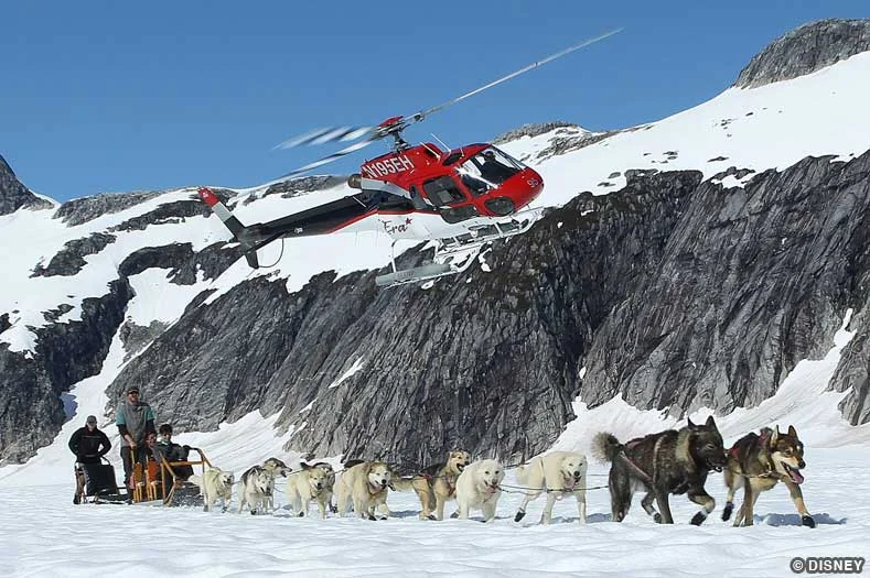 A team of sled dogs runs along a snowy mountainscape as a helicopter buzzes overhead
