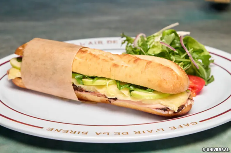 A crusty baguette, split and containing black pepper, arugula, apples, Brie, mustard butter, and apple butter. A side salad is also on the plate.